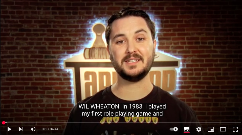 Wil Wheaton's head and shoulders in front of the Tabletop logo on a red brick wall. He is a guy in his thirties with brown, short hair and beard. He is wearing a brown t-shirt with "the Guild" on it. The closed caption on the screen reads: "WIL WHEATON: In 1983, I played my first role playing game and"