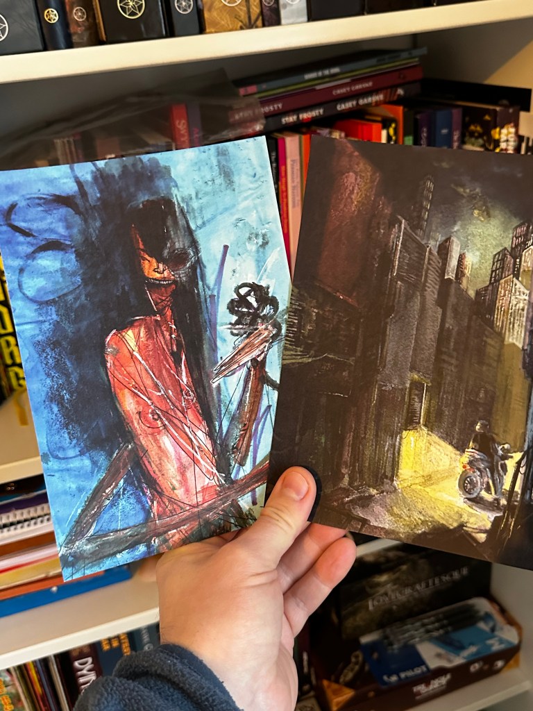 Two art prints: On the left, a spiky, crimson femme creature with wide open mouth, long black hair against a blue background. On the right, a person on a motorbike stopped in a pool of yellow light from a doorway in the backstreet of a city. The cityscape rises above and behind.