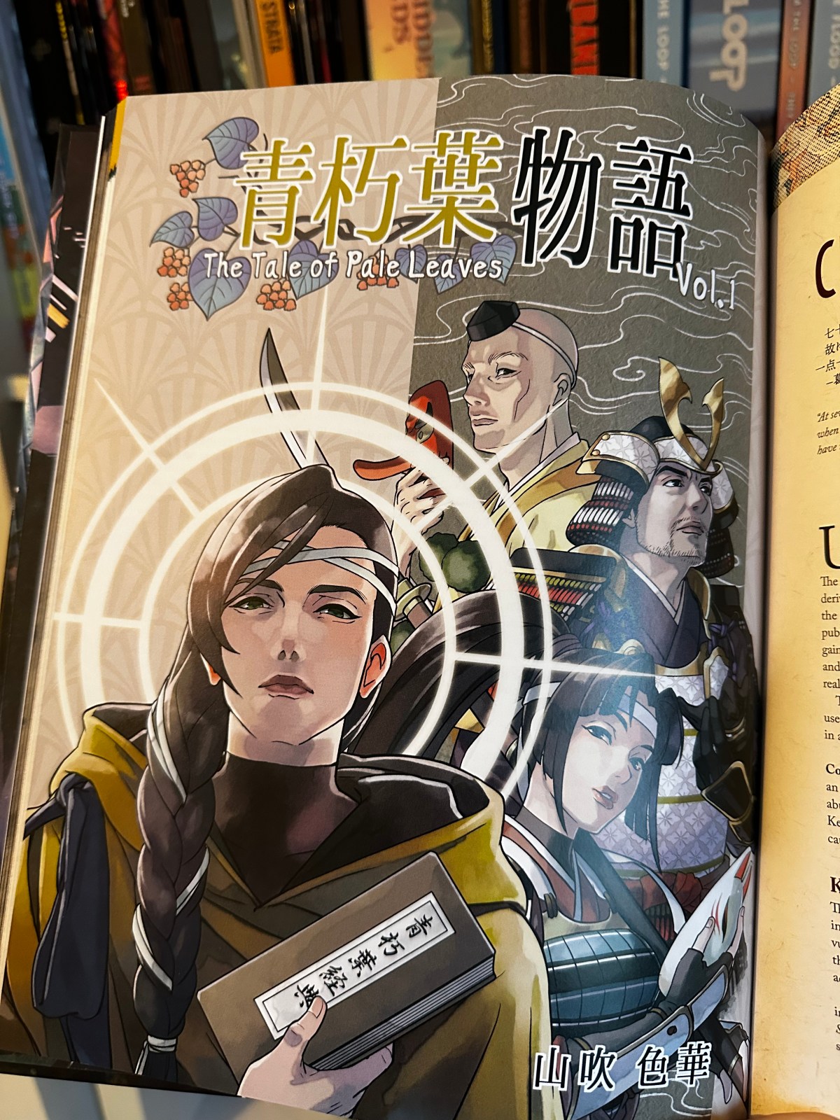 The cover of Aokuchiba, the Tale of Pale Leaves by Yamabuki Iroha. Its a manga style artwork in colour featuring a handsome prince, holding the sutra and wearing yellow robes and backed by a cast of secondary characters including a samurai and a woman holding a kitsune mask