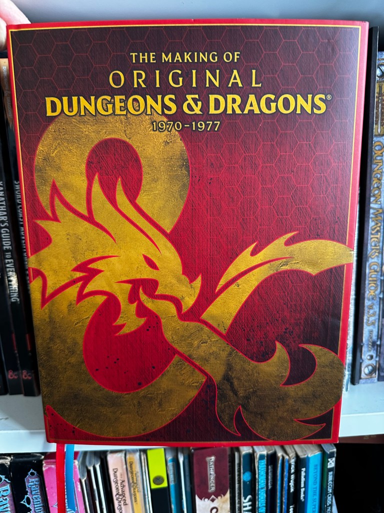 The cover of the Making of the Original Dungeons and Dragons. It has a red cover with a big gold ampersand on it.