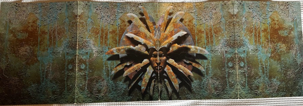 The outside of the Planescape DM screen. The visage of the Lady of Pain fis the centre-piece. It is the face of a severe looking woman surrounded by a head-dress of blades. It looks like its made of verdigris-tinged copper.