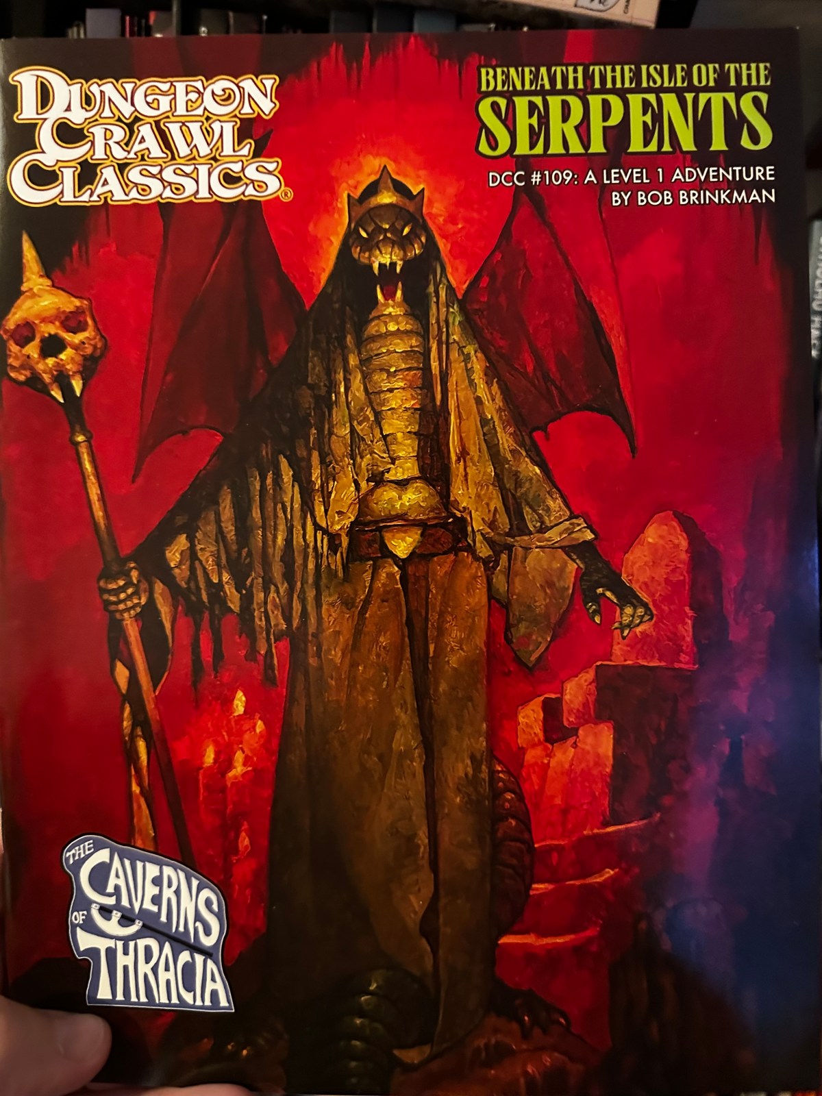 The cover of DCC # 109 Beneath the Isle of the Serpents by Bob Brinkman. The cover is very red and depicts a winged and crowned serpent person with a skull staff standing before a throne