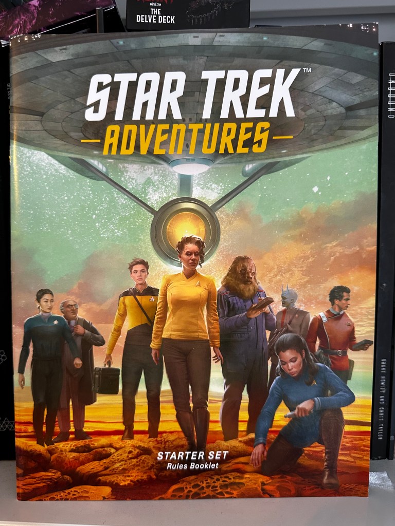 Star Trek Adventures Starter Set Rules Booklet Cover. It features a variety of star fleet personnel from different eras on the surface of a desert planet with a star fleet ship looming above them.